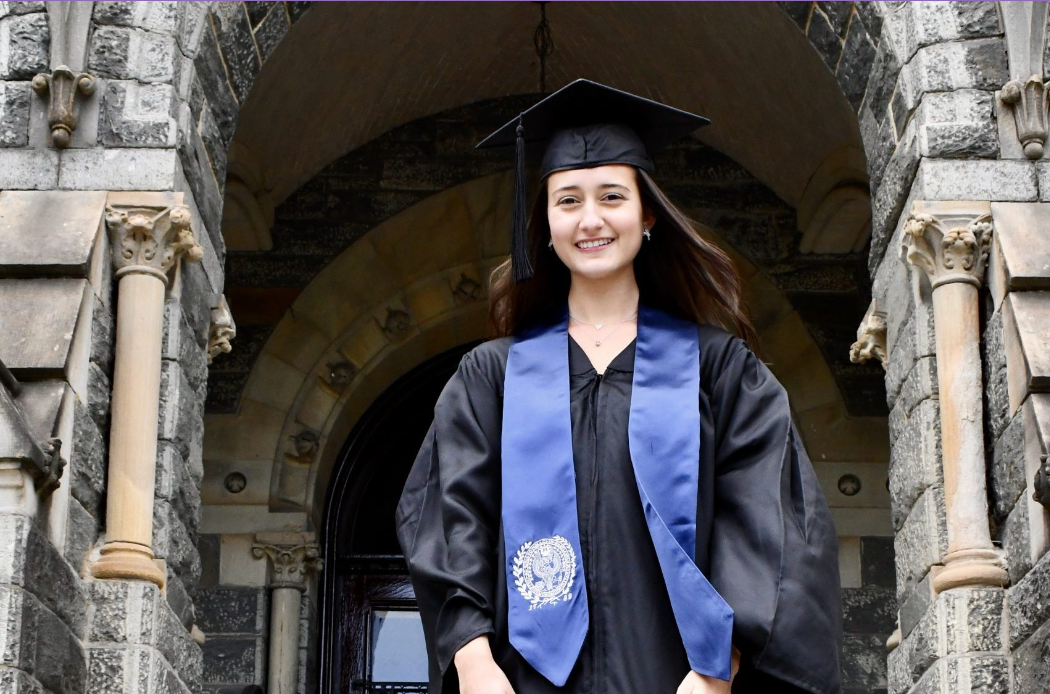 Photo of Ana Lucía Marín Jaramillo in a black graduation cap and grown and a blue sash, in front of a building