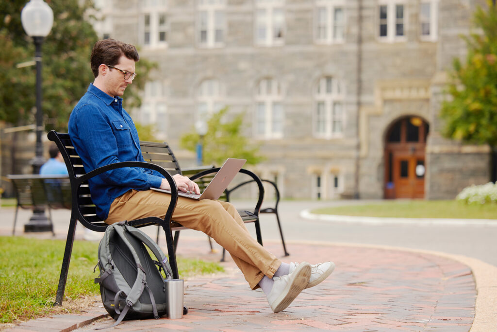 MBA Student sitting on bench on Healy Lawn working on laptop for their online masters program