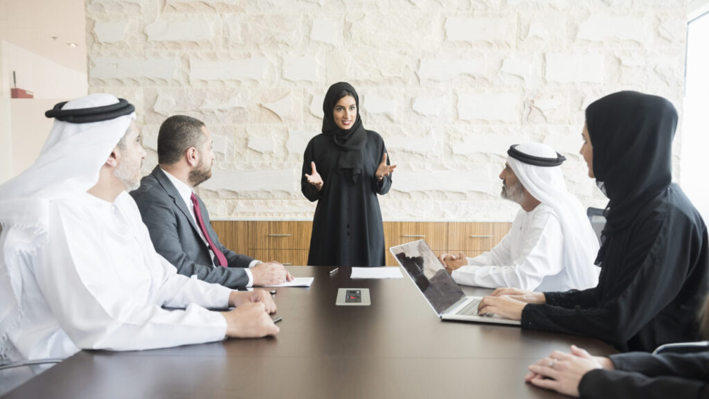A photo of young and smiling Arab businesswoman gesturing while giving presentation. Emirati business people are wearing traditional clothes of those and abaya. Multi-ethnic colleagues are listening to her very carefully. All are in brightly lit office. EMBA Dubai, Executive MBA Dubai.