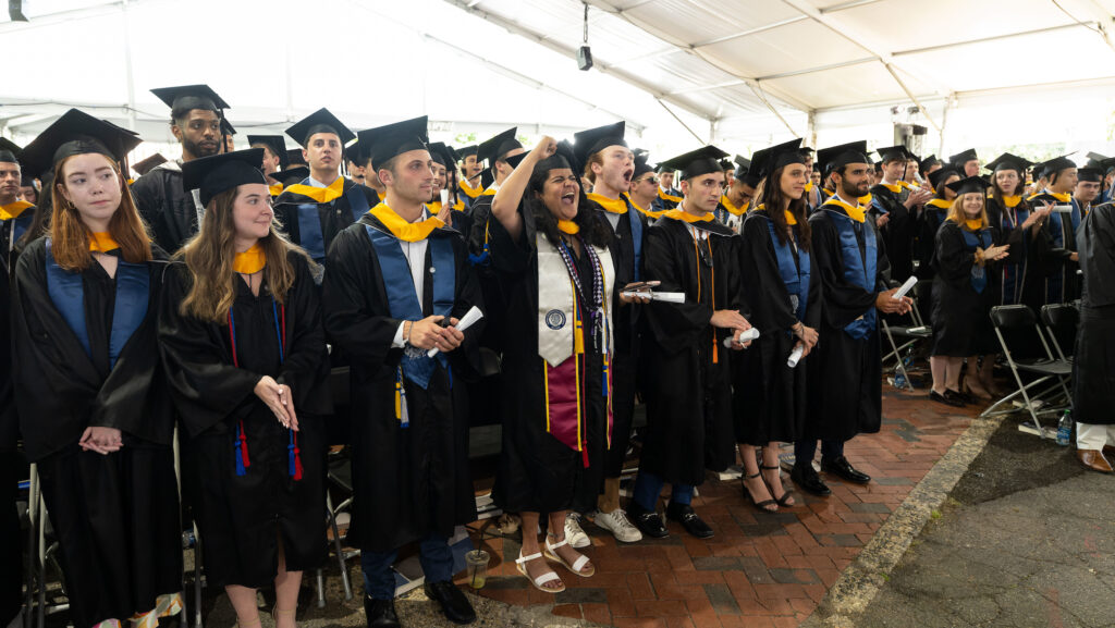 Undergraduate students standing at their graduation listening to speaker