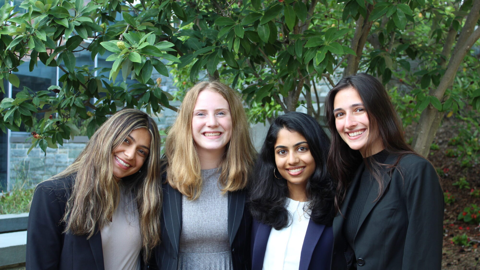 4 women undergraduates wearing blazers smiling outside in front of a tree