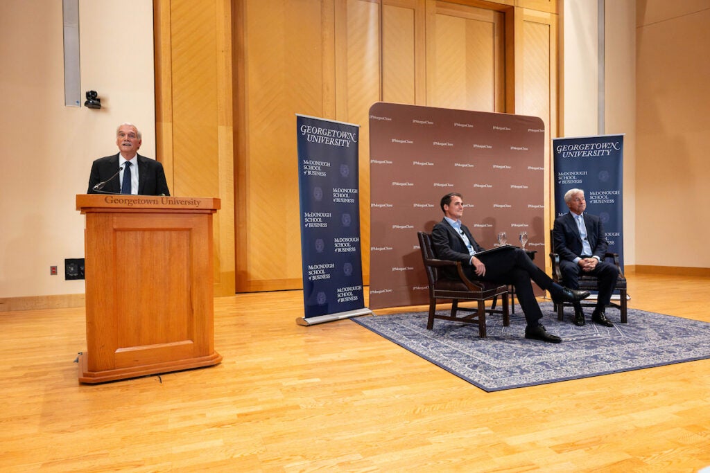 Georgetown President Robert Groves stands behind a lectern on stage with Alberto Rossi and Jamie Dimon seated in front of banners