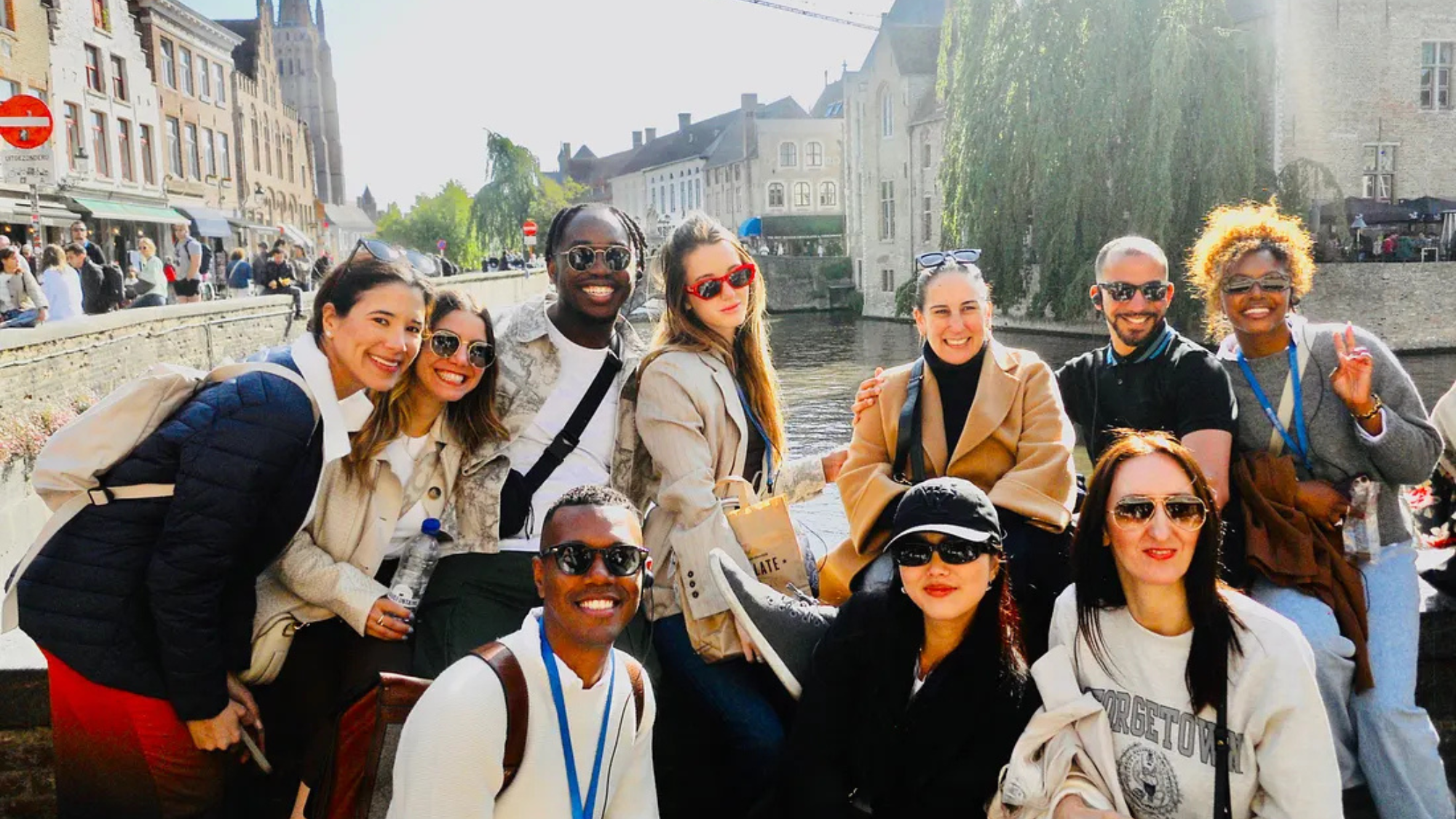 Students in various color shirts, hats and sunglasses pose in front of a bridge in Brussels.