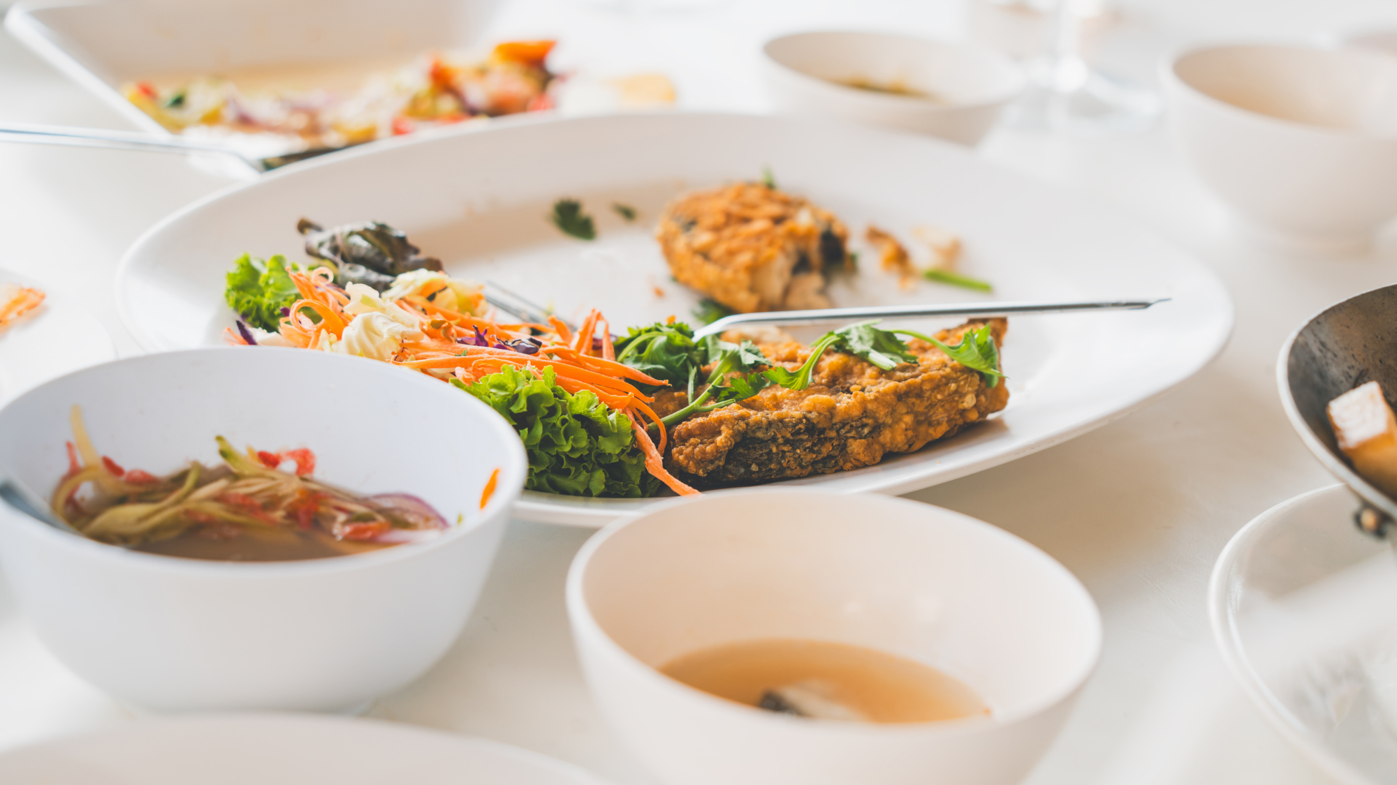Plates and bowls with colorful foods that are half eaten and silverware left on a white linen-covered table