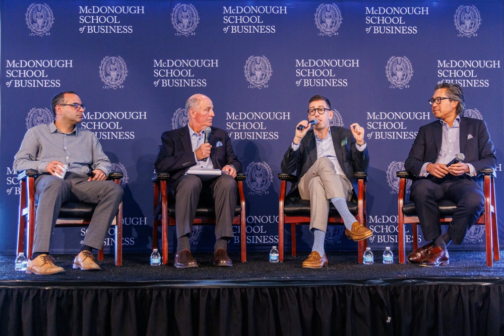 Four men sit on stage in front of Georgetown McDonough step-and-repeat for Business of Health Summit