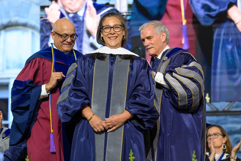 Paul Almeida (L) and Interim President Robert Groves (R) put the honorary doctorate hood on Essya Hanachi (B’91)